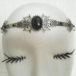 May include: A silver headband with a black gemstone and intricate filigree design. The headband is on a white mannequin head.