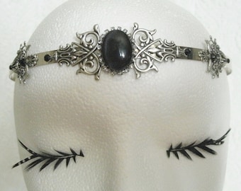 Puede incluir: Una diadema plateada con una piedra preciosa negra y un diseño filigranado intrincado. La diadema está en una cabeza de maniquí blanca.