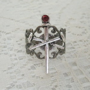 May include: A silver ring with a red gem on top of a sword design. The ring is ornate and has a filigree design.