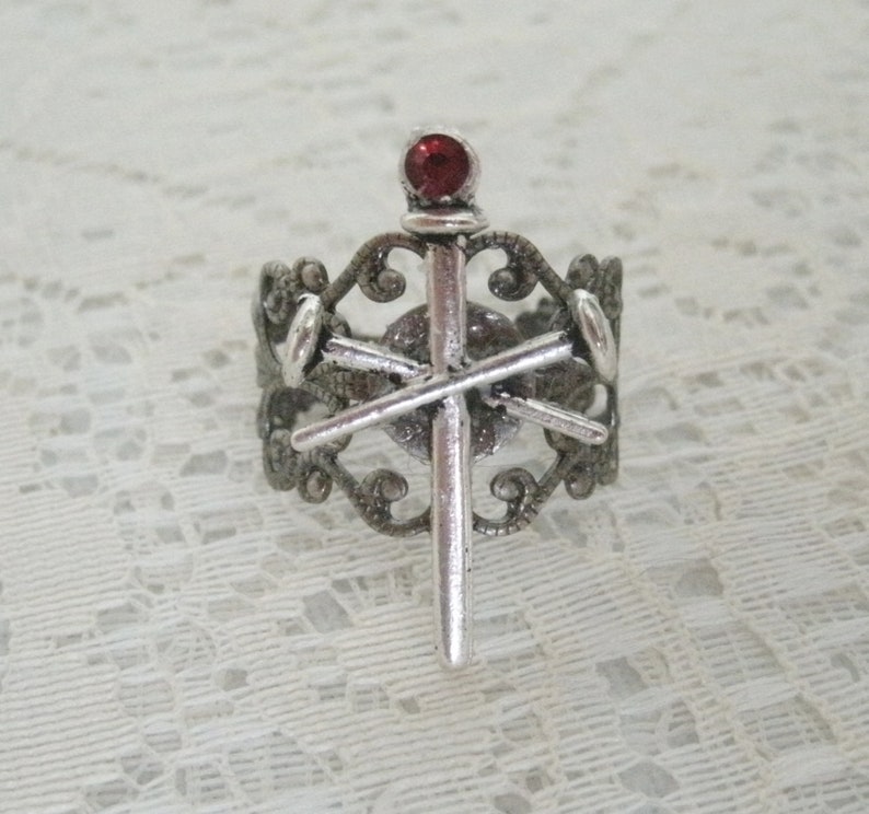 May include: A silver ring with a red gem on top of a sword design. The ring is ornate and has a filigree design.
