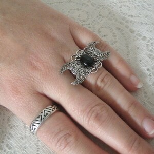 May include: A silver ring with a black stone and a triple moon design. The ring is on a finger with a second silver ring with a geometric design.