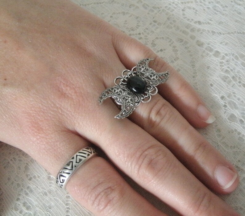 May include: A silver ring with a black stone and a triple moon design. The ring is on a finger with a second silver ring with a geometric design.