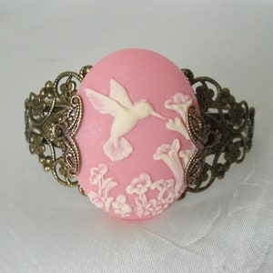 May include: A pink cameo bracelet with a white hummingbird and floral design. The bracelet is made of metal with a filigree design.