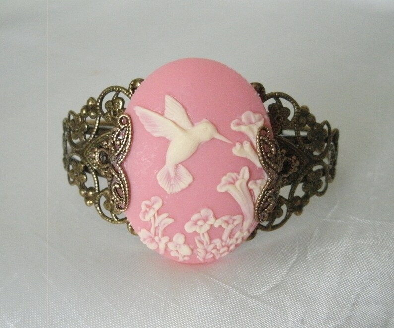 May include: A pink cameo bracelet with a white hummingbird and floral design. The bracelet is made of metal with a filigree design.