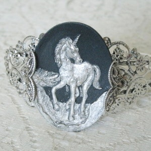 May include: A silver bracelet with a black cameo featuring a silver unicorn. The cameo is surrounded by a silver filigree design.