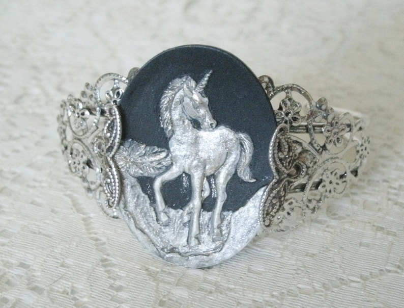 May include: A silver bracelet with a black cameo featuring a silver unicorn. The cameo is surrounded by a silver filigree design.