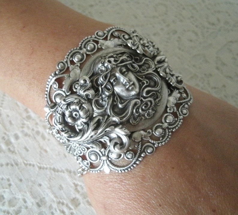 K&ouml;nnte beinhalten: Silberarmband mit einem verzierten Design, das ein Frauen Gesicht zeigt, umgeben von Blumen und Bl&auml;ttern.