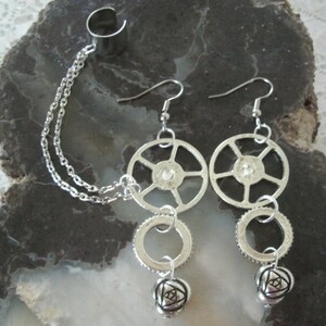 May include: Silver ear cuff with a chain connecting to a silver dangle earring. The earring features a wheel-shaped charm, a ring charm, and a rose-shaped charm.
