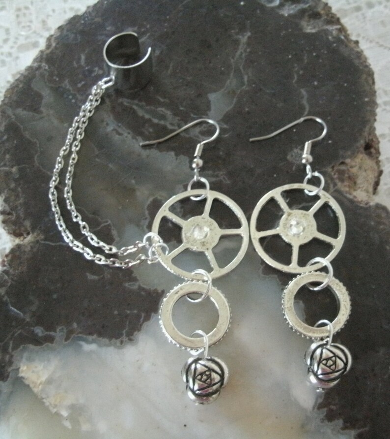 May include: Silver ear cuff with a chain connecting to a silver dangle earring. The earring features a wheel-shaped charm, a ring charm, and a rose-shaped charm.