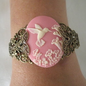 May include: A pink cameo bracelet with a white hummingbird and floral design. The bracelet is made of metal with a filigree design.