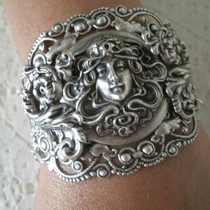 K&ouml;nnte beinhalten: Silberarmband mit einem verzierten Design, das ein Frauen Gesicht zeigt, umgeben von Blumen und Bl&auml;ttern.