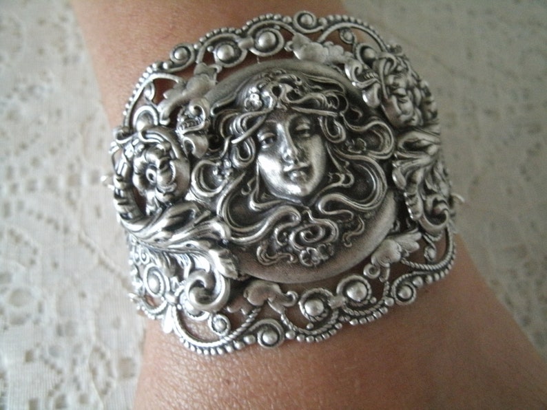 K&ouml;nnte beinhalten: Silberarmband mit einem verzierten Design, das ein Frauen Gesicht zeigt, umgeben von Blumen und Bl&auml;ttern.