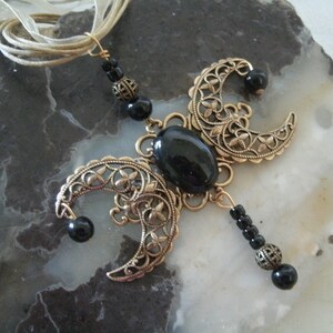 Puede incluir: Collar artesanal con un colgante de metal color bronce. El colgante presenta formas de media luna con calados florales, una piedra negra ovalada central y cuentas negras. Una cinta color crema sirve como cadena del collar.