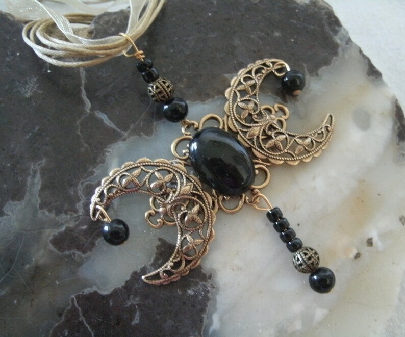 Puede incluir: Collar artesanal con un colgante de metal color bronce. El colgante presenta formas de media luna con calados florales, una piedra negra ovalada central y cuentas negras. Una cinta color crema sirve como cadena del collar.