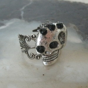 May include: A silver skull ring with black rhinestone accents. The ring has a detailed design with a filigree band.