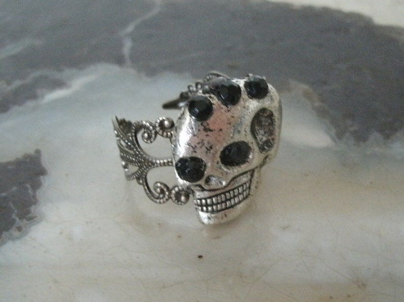 May include: A silver skull ring with black rhinestone accents. The ring has a detailed design with a filigree band.