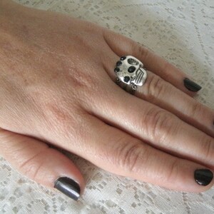 May include: A silver skull ring with black accents. The ring is worn on the index finger of a hand with black nail polish.