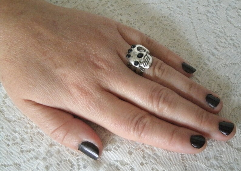 May include: A silver skull ring with black accents. The ring is worn on the index finger of a hand with black nail polish.