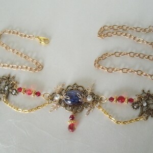 Op de afbeelding: Een gouden ketting met een blauwe en witte cameo hanger, rode kralen en gouden filigraan accenten.