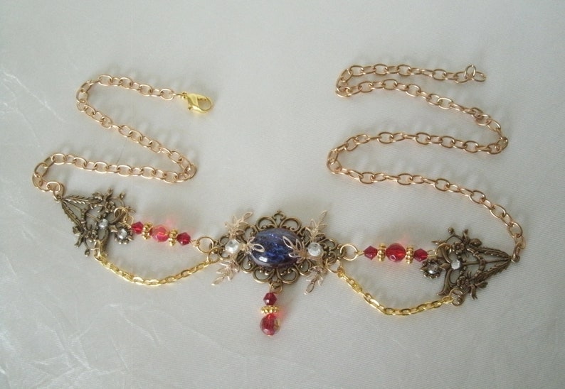 Op de afbeelding: Een gouden ketting met een blauwe en witte cameo hanger, rode kralen en gouden filigraan accenten.