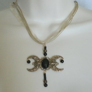 Puede incluir: Collar con colgante plateado con un dise&ntilde;o de triple luna y una gran piedra ovalada negra. El colgante cuelga de una cinta transparente de varios hilos. Estilo g&oacute;tico.