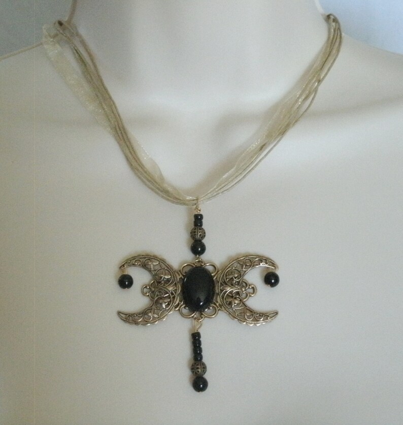 Puede incluir: Collar con colgante plateado con un dise&ntilde;o de triple luna y una gran piedra ovalada negra. El colgante cuelga de una cinta transparente de varios hilos. Estilo g&oacute;tico.