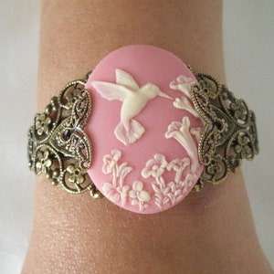 May include: A pink cameo bracelet with a white hummingbird and floral design. The bracelet is made of metal with an ornate design.