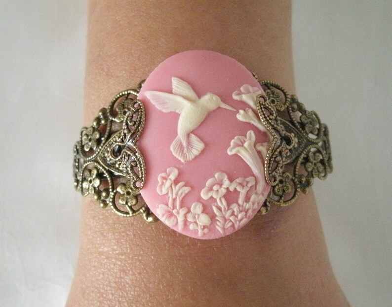 May include: A pink cameo bracelet with a white hummingbird and floral design. The bracelet is made of metal with an ornate design.