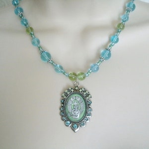 May include: A beaded necklace featuring a pendant with a light green background and a silver-colored frame. The necklace has light blue and green beads. The pendant has a detailed illustration of a figure with wings.