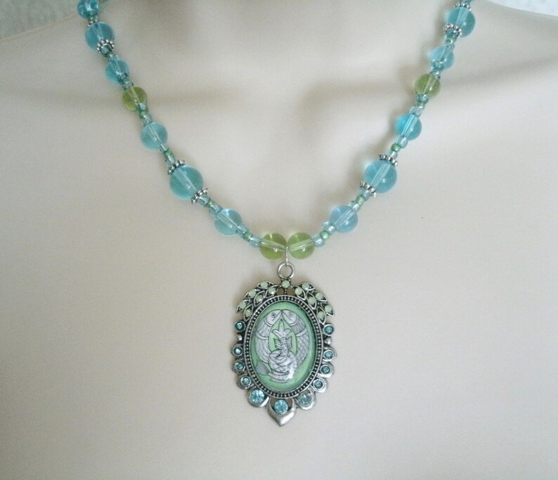 May include: A beaded necklace featuring a pendant with a light green background and a silver-colored frame. The necklace has light blue and green beads. The pendant has a detailed illustration of a figure with wings.