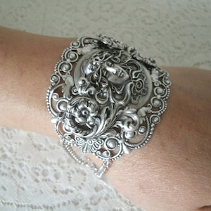 K&ouml;nnte beinhalten: Silberarmband mit einem verzierten Design, das im Zentrum ein Frauen Gesicht zeigt. Das Armband hat ein Kettendetail und ist aus Metall.