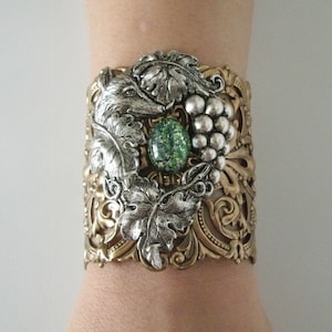 May include: A silver and gold cuff bracelet with a detailed design of leaves, grapes, and a fox. The bracelet features a green gemstone in the center.
