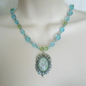 May include: A beaded necklace featuring a pendant with a light green background and a silver-colored frame. The necklace has light blue and green beads. The pendant has a detailed design with a light green background and a silver-colored frame.