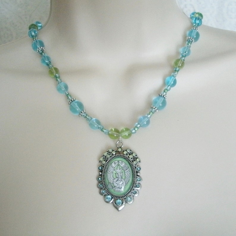 May include: A beaded necklace featuring a pendant with a light green background and a silver-colored frame. The necklace has light blue and green beads. The pendant has a detailed design with a light green background and a silver-colored frame.