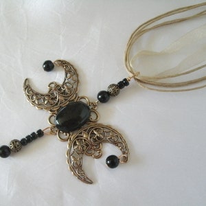 Puede incluir: Un collar con colgante de color bronce con una piedra negra ovalada central y formas de media luna con recortes florales. Cuentas negras y una cinta beige completan el dise&ntilde;o. El collar tiene una est&eacute;tica vintage y g&oacute;tica.
