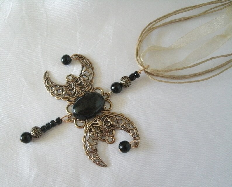 Puede incluir: Un collar con colgante de color bronce con una piedra negra ovalada central y formas de media luna con recortes florales. Cuentas negras y una cinta beige completan el dise&ntilde;o. El collar tiene una est&eacute;tica vintage y g&oacute;tica.