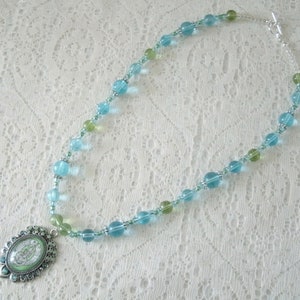 May include: A beaded necklace featuring a pendant with a green and white design. The necklace has a mix of light blue and green beads, with silver-colored accents. The necklace is displayed on a white lace surface.