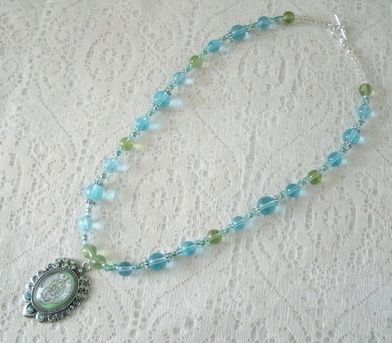 May include: A beaded necklace featuring a pendant with a green and white design. The necklace has a mix of light blue and green beads, with silver-colored accents. The necklace is displayed on a white lace surface.