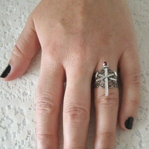 May include: A silver ring with a red stone and a silver cross design.