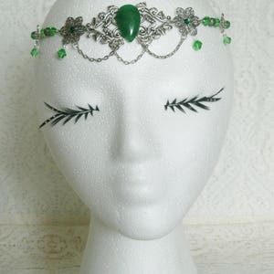 Puede incluir: Una diadema plateada con una piedra preciosa verde y cuentas de vidrio verdes. La diadema est&aacute; en una cabeza de poliestireno blanco.