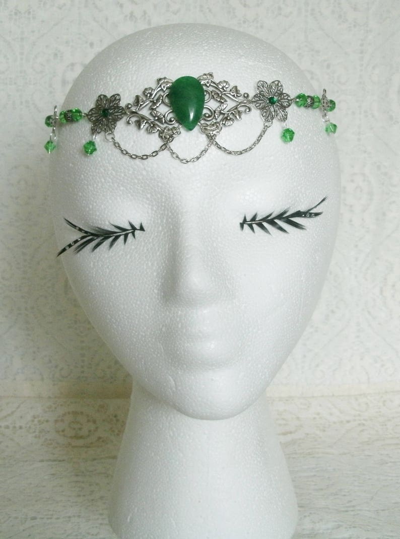 Puede incluir: Una diadema plateada con una piedra preciosa verde y cuentas de vidrio verdes. La diadema est&aacute; en una cabeza de poliestireno blanco.