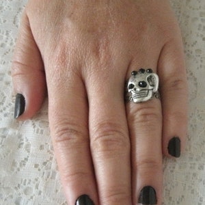 May include: A silver skull ring with black gem accents. The ring is worn on the middle finger of a hand with black nail polish.