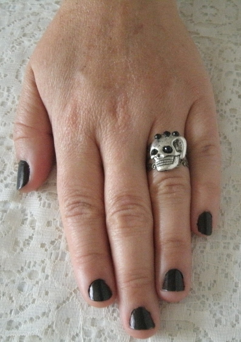 May include: A silver skull ring with black gem accents. The ring is worn on the middle finger of a hand with black nail polish.