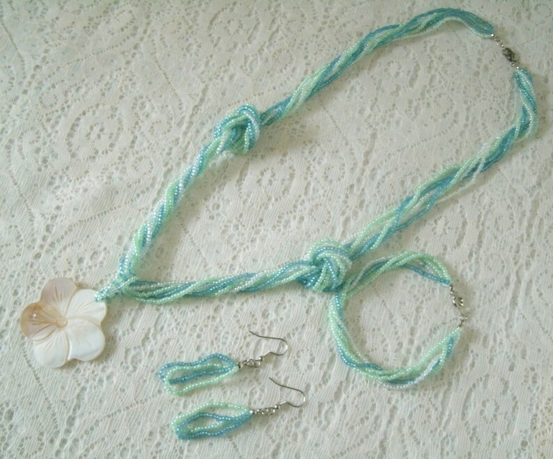 May include: A set of blue and white beaded jewelry, including a necklace, bracelet, and earrings. The necklace features a large, white shell flower pendant.