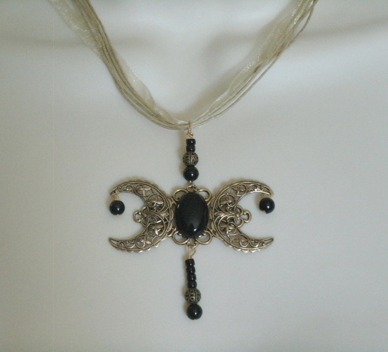 Puede incluir: Collar con colgante plateado con un dise&ntilde;o de triple luna y una gran piedra negra ovalada en el centro. Las medias lunas tienen intrincados patrones de filigrana y cuentas negras. La cadena es delicada, transparente y similar a una cinta.