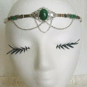 May include: Silver headband with a green gemstone and smaller green stones on the sides. The headband has a delicate chain design.