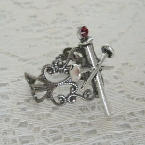 May include: Silver ring with a delicate filigree design. A silver sword with a red gemstone at the top is attached to the ring.