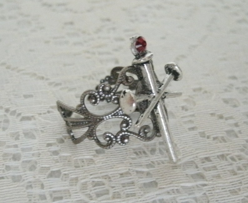 May include: Silver ring with a delicate filigree design. A silver sword with a red gemstone at the top is attached to the ring.