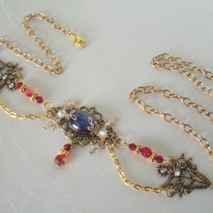 Op de afbeelding: Een gouden ketting met een grote blauwe en witte edelsteen hanger. De hanger is omgeven door ingewikkelde gouden filigraan en kleine rode en witte kralen. De ketting is versierd met kleine rode kralen.