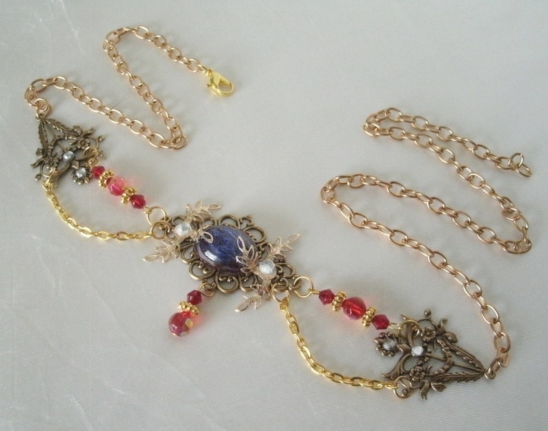 Op de afbeelding: Een gouden ketting met een grote blauwe en witte edelsteen hanger. De hanger is omgeven door ingewikkelde gouden filigraan en kleine rode en witte kralen. De ketting is versierd met kleine rode kralen.